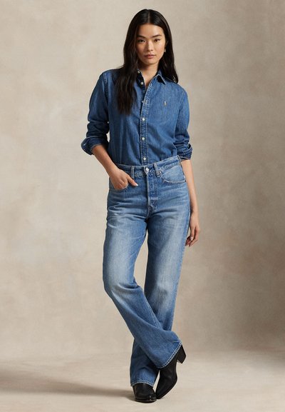 Femeie în picioare cu o mână în buzunar, purtând o cămașă din denim cu mâneci lungi, blugi de culoare albastru deschis și cizme negre cu toc.