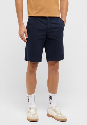 Mann trägt marineblaue Shorts, weiße Socken mit schwarzem Text und beige-weiße Turnschuhe, steht mit den Händen in den Taschen.
