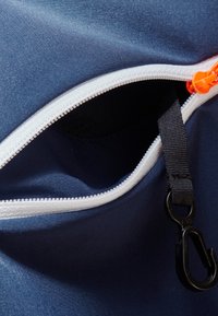 Rogaty niebieski worek materiałowy w rogu z białym zamkiem błyskawicznym, pomarańczowym uchwytem i czarnym klipsem. Gładka tekstura, minimalistyczny design, otwarty od góry.