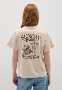 T-shirt en coton beige avec un graphique imprimé marron représentant une tasse de café, un croissant et une cafetière, avec le texte "SUNDAY Brunch" et "Morning Club".