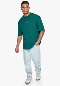 Camiseta oversized de color verde azulado con cuello redondo y un pequeño parche de logo, combinada con unos pantalones vaqueros holgados de color azul claro y zapatillas blancas.
