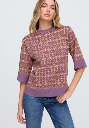 Frau trägt ein violett gemustertes Strickoberteil mit dreiviertellangen Ärmeln und blaue Jeans, steht mit einer Hand auf der Hüfte vor einem schlichten Hintergrund.