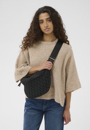 Jauna moteris su ilgais garbanotais plaukais dėvi smėlio spalvos megztinį ir mėlynas džinsus, laikydama juodą siuvinėtą per petį nešiojamą rankinę su plačia diržine juosta per krūtinę.