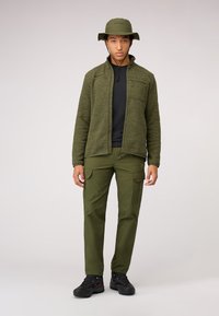 Giacca in pile verde con tasche con zip, t-shirt nera, pantaloni cargo verdi e scarpe da escursionismo nere. Include un cappello a secchiello verde coordinato.