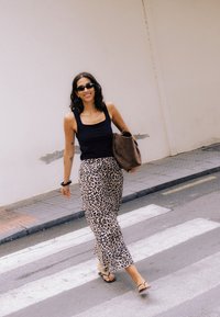 Frau mit Sonnenbrille und Leoparden-Print-Rock geht über eine Straße, trägt eine braune Tasche unter dem Arm und lächelt.