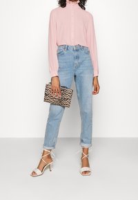 Blouse rose clair à manches longues avec boutons sur le devant, col montant, jean taille haute bleu, et sandales blanches à talons avec brides. Pochette imprimé léopard.