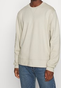 Homme portant un sweat-shirt beige à manches longues avec col rond et un jean bleu, les mains détendues le long du corps, fond blanc uni.