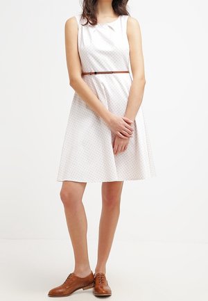 Femme portant une robe blanche sans manches, longueur genou, avec un motif subtil, une fine ceinture marron et des chaussures en cuir marron à lacets, debout les mains jointes.