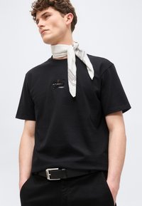 Νέος άνδρας με μαύρο T-shirt με διακοσμητική ετικέτα λογότυπου, μαύρη ζώνη και λευκό κασκόλ με σχέδια στη δεξιά πλευρά του λαιμού.