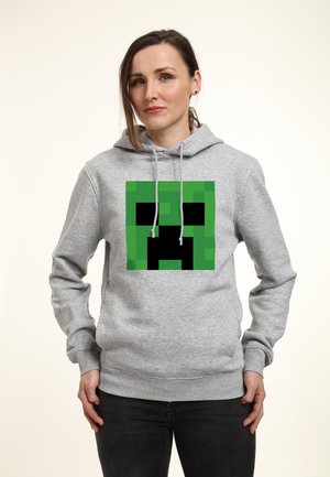 Kobieta w szarej bluzie z kapturem, z dużą zieloną i czarną pikselową twarzą Creepera z Minecrafta na przodzie, stoi z rękami w kieszeniach.