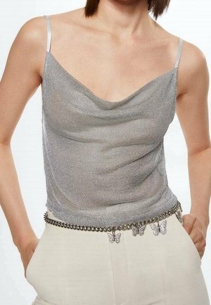 Srebrny metaliczny top bez rękawów z cienkimi ramiączkami, zestawiony z kremowymi spodniami z wysokim stanem oraz łańcuszkowym paskiem ozdobionym zawieszkami w kształcie motyli.