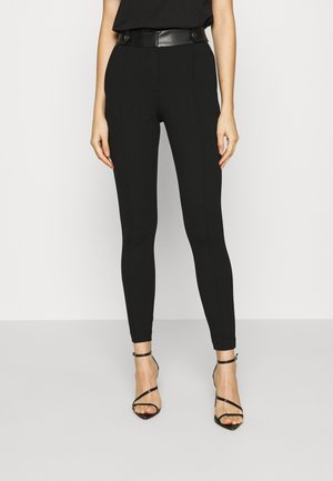 Schwarze, körpernahe Hosen mit hoher Taille und glänzendem schwarzen Gürtel. Glattes Material und elegantes Design. Kombiniert mit schwarzen Riemchensandalen.