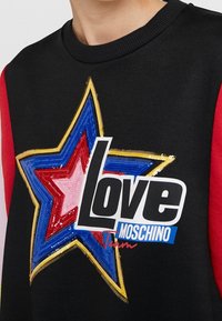 Mustat hupparit, joissa on suuri monivärinen tähden kuvio koristeltuna paljeteilla. Sana "Rakkaus" valkoisella on näkyvästi esillä keskellä.