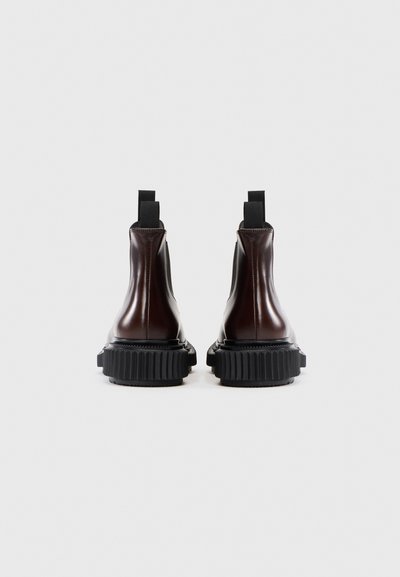 Botine Chelsea din piele lucioasă maro, cu panouri elastice pe laterale, tabul de tragere și tălpi groase din cauciuc negru cu textură nervurată.