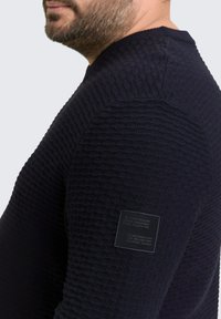 Marineblauwe gebreide trui met een gestructureerd patroon, ronde halslijn en een klein merklabel op de linker schouder.