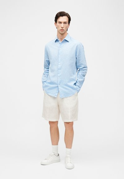 Άνδρας στέκεται με τα χέρια στις τσέπες, φορώντας ανοιχτό μπλε πουκάμισο με μακριά μανίκια και κουμπιά, λευκά σορτς μέχρι το γόνατο, λευκές κάλτσες και λευκά αθλητικά παπούτσια.