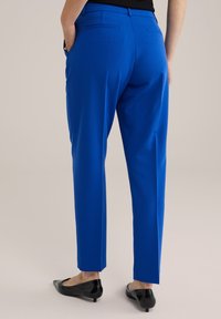 Mėlynos pritaikytos kelnės su lygia tekstūra, turinčios šonines kišenes ir liesą siluetą, derinamos su juodomis butais prieš neutralų foną.