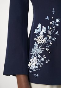 Manche et torse bleu marine d'un vêtement avec broderie florale bleu clair et regroupements de perles blanches ainsi que des embellissements en perles transparentes.