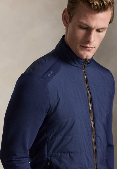 Hombre con chaqueta acolchada azul marino con cremallera y logo RLX en el hombro, mirando hacia abajo sobre un fondo liso beige.