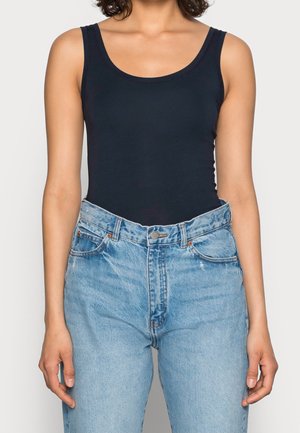 Mujer con camiseta negra sin mangas metida en unos vaqueros de mezclilla azul claro de cintura alta con botones delanteros y bolsillos.