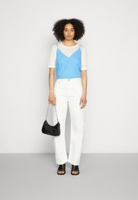 Camisola azul sobre uma t-shirt branca, acompanhada de jeans brancos de perna larga. Modelo segura uma mala preta e usa sandálias de salto pretas.