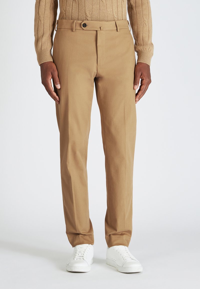 Pantalones de algodón beige con un diseño de pierna recta, que cuentan con un cierre de botón y pliegues sutiles, combinados con zapatillas blancas.