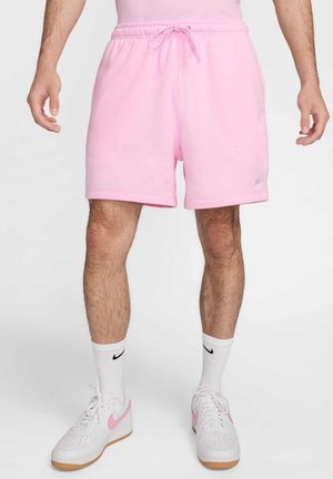 Homme portant un short rose clair avec cordon, chaussettes blanches Nike et baskets blanches avec un swoosh rose, debout devant un fond uni.
