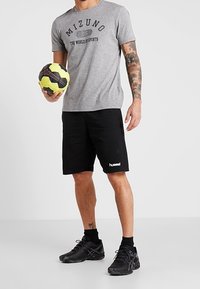 Mężczyzna w szarej koszulce Mizuno i czarnych szortach Hummel trzymający żółtą i czarną piłkę ręczną, stojący na jednolitym tle.