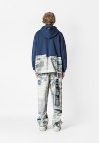 Sudadera navy con un panel inferior de denim texturizado y desgastado, combinada con pantalones de un azul claro y con estampado que presentan una etiqueta vertical y suelas blancas gruesas.
