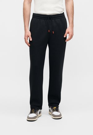 Mann trägt schwarze Jogginghose mit Kordelzug, weiße Sneaker mit schwarzen und grauen Akzenten sowie ein weißes T-Shirt und steht vor einem einfarbigen Hintergrund.