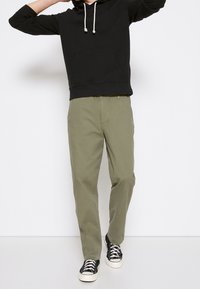 Sweat à capuche noir avec poche avant et cordon, associé à un pantalon vert olive clair. Des baskets noires et blanches complètent la tenue.
