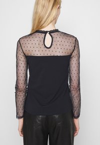 Svart topp med genomskinliga långärmar med prickar, rund halsringning och en nyckelhålsdetalj bak. Textuerad tyg kontrasterar med den släta kroppen.