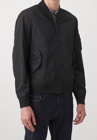 Giubbotto bomber nero con chiusura a zip, polsini a coste e dettaglio tasca ovale sul lato sinistro. Realizzato in tessuto leggero e testurizzato.