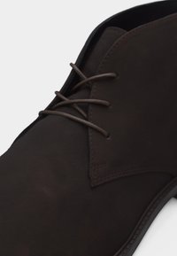 Σκούρο καφέ δερμάτινο παπούτσι επίσημο με στρογγυλεμένη μύτη, ελάχιστη ραφή και λεπτά κορδόνια ίδιου χρώματος δεμένα με έναν απλό κόμπο.