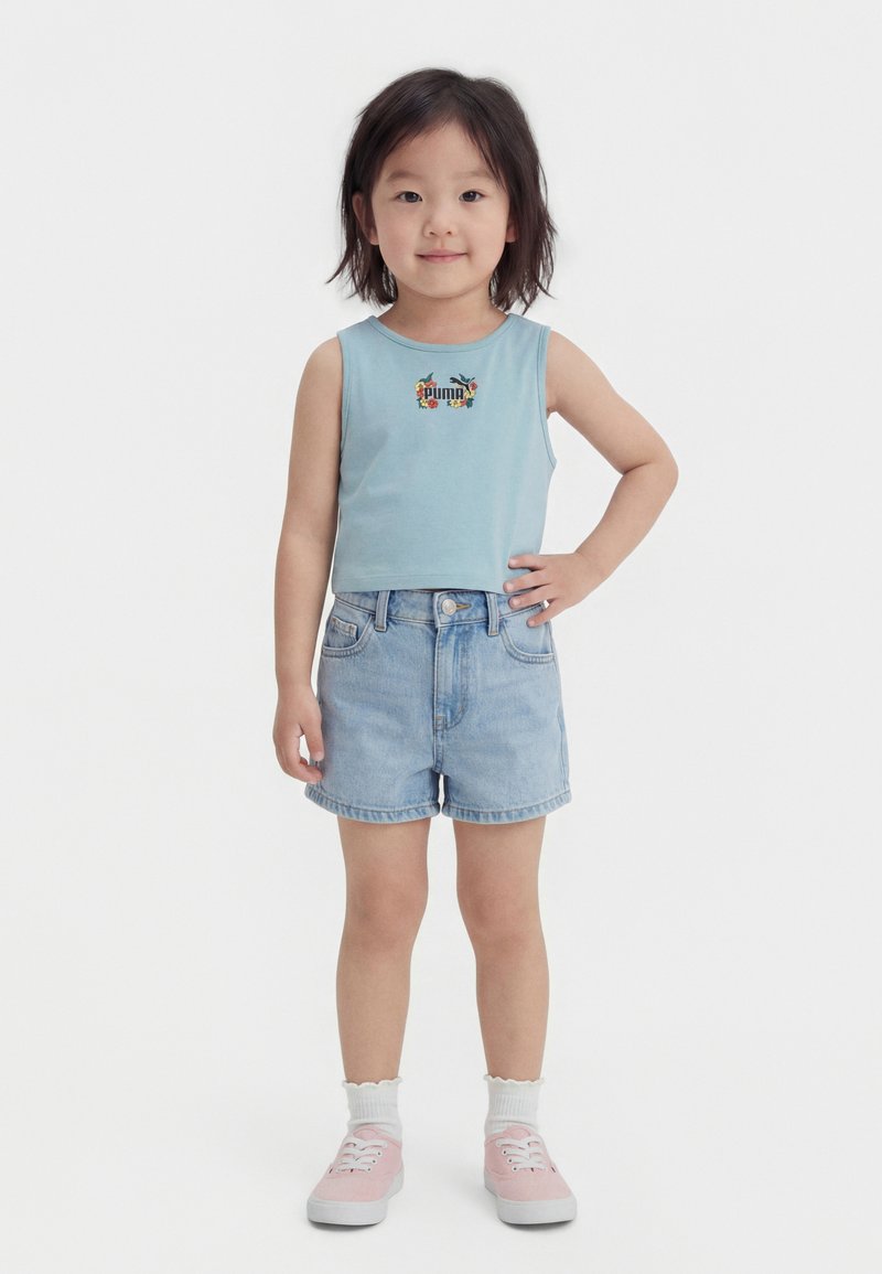 Unge barn står med hånden på hoften iført lyseblå Puma tanktop, denimshorts, hvide sokker og lyserøde sneakers på hvid baggrund.