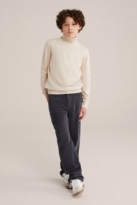 Bež vestica s turtleneck izradom, teksturirana tkanina; uparena s tamnosivim baršunastim hlačama. Model nosi bijele tenisice s svijetlosmeškanim akcentima.