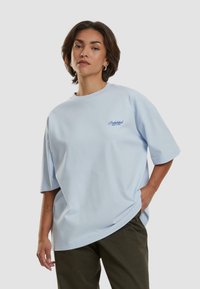 Lys blå oversized t-skjorte i myk bomull med rund hals og korte ermer; har liten blå brodert tekst på brystet.