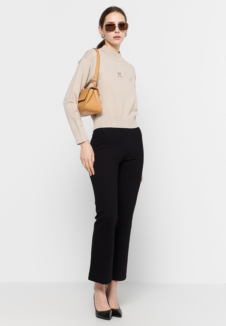 Maglione beige a maglia con un effetto metallico, pantaloni neri sartoriali alla caviglia e una borsa in pelle color cuoio. Modella indossa occhiali da sole e tacchi.