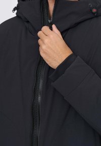 Hånd trækker lynlås op på sort hættejakke med justerbar snor og tekstureret vandtæt lynlåsdetalje.