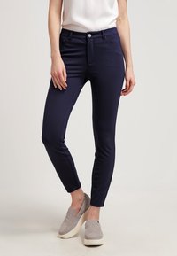 Marina stretchbyxor med slim fit, medelhög midja, framfickor och en mjuk yta. Matchat med gråa skor utan snören.