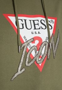 Olivově zelená mikina s kapucí s velkým červenobílým trojúhelníkovým logem "GUESS" v černém textu, doplněná třpytivými nápisy z kamínků "Icon."