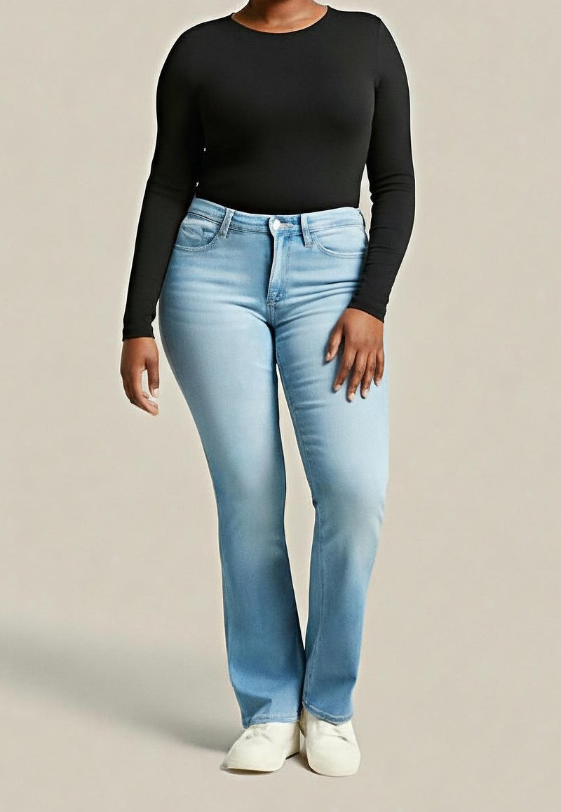 Persona con jeans acampanados azul claro, camiseta negra ajustada de manga larga y zapatos blancos, de pie frente a un fondo liso.