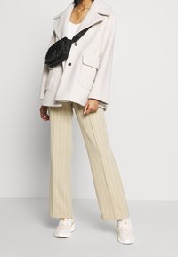 Cappotto in pile beige oversize con grandi tasche, abbinato a pantaloni a palazzo a righe beige chiaro e scarpe da ginnastica bianche. Borsa a tracolla nera.