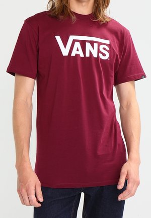 Person, der ein kastanienbraunes Vans-T-Shirt mit großem weißen Vans-Logo auf der Brust trägt, kombiniert mit dunkelblauen Jeans.