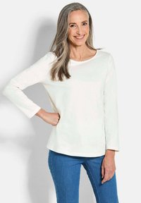 Witte lange mouwen shirt met een ronde hals, gladde textuur, eenvoudige ontwerp, gecombineerd met blauwe strakke jeans. Model staat met de hand op de heup.