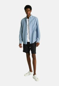 Chemise à manches longues bleu clair avec des boutons, portée par-dessus un t-shirt blanc, associée à un short noir et des sneakers blanches. Tissu en coton, coupe décontractée.