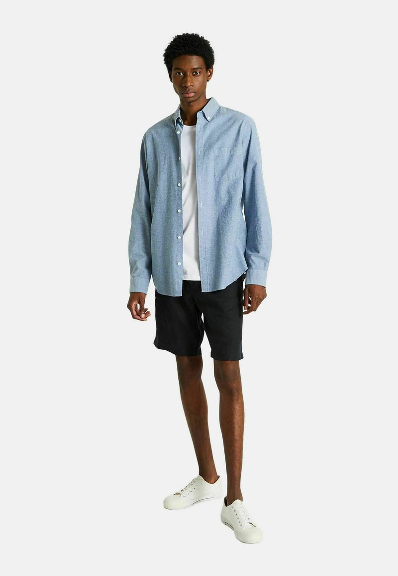 Chemise à manches longues bleu clair avec des boutons, portée par-dessus un t-shirt blanc, associée à un short noir et des sneakers blanches. Tissu en coton, coupe décontractée.