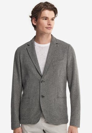 Ung mand iført en grå to-knaps blazer over en hvid T-shirt, kiggende let mod højre mod en ensfarvet baggrund.