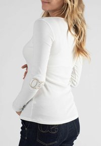 Wit ribbed shirt met lange mouwen, featuring een getailleerd ontwerp en gouden accenten op de ellebogen. Gecombineerd met donkerblauwe jeans, wat een casual uitstraling geeft.