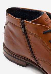 Bottine en cuir marron avec une fermeture éclair latérale et une doublure en tissu noir. Accents cousus avec une semelle en caoutchouc et une finition texturée.